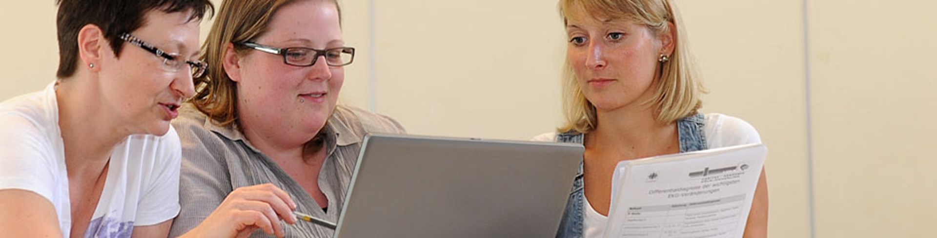Drei Frauen sitzen an einem Tisch und schauen auf einen Laptop. Eine Frau zeigt auf den Bildschirm, während die andere eine Unterlage in der Hand hält.